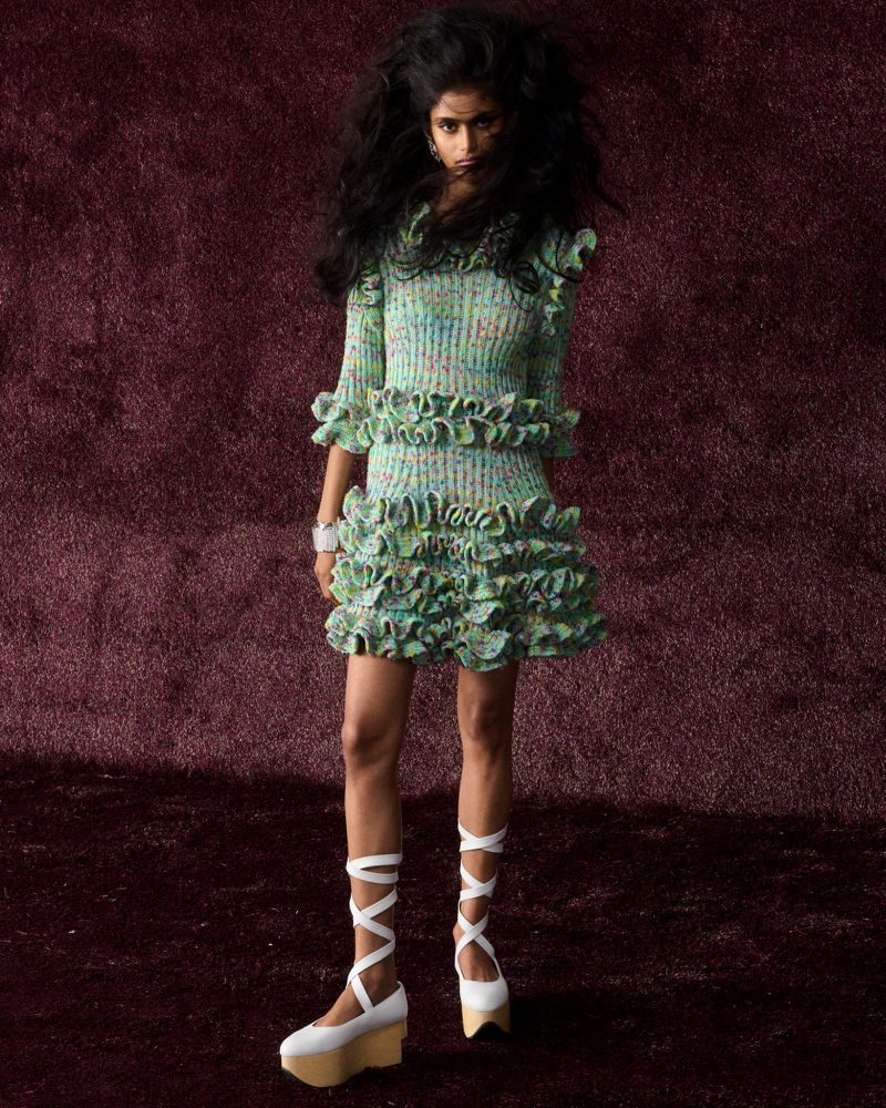 Fashion shot of woman with afro, wearing a knit dress with ruffles and ribbon platform shoes in a photo studio with a brown backdrop.