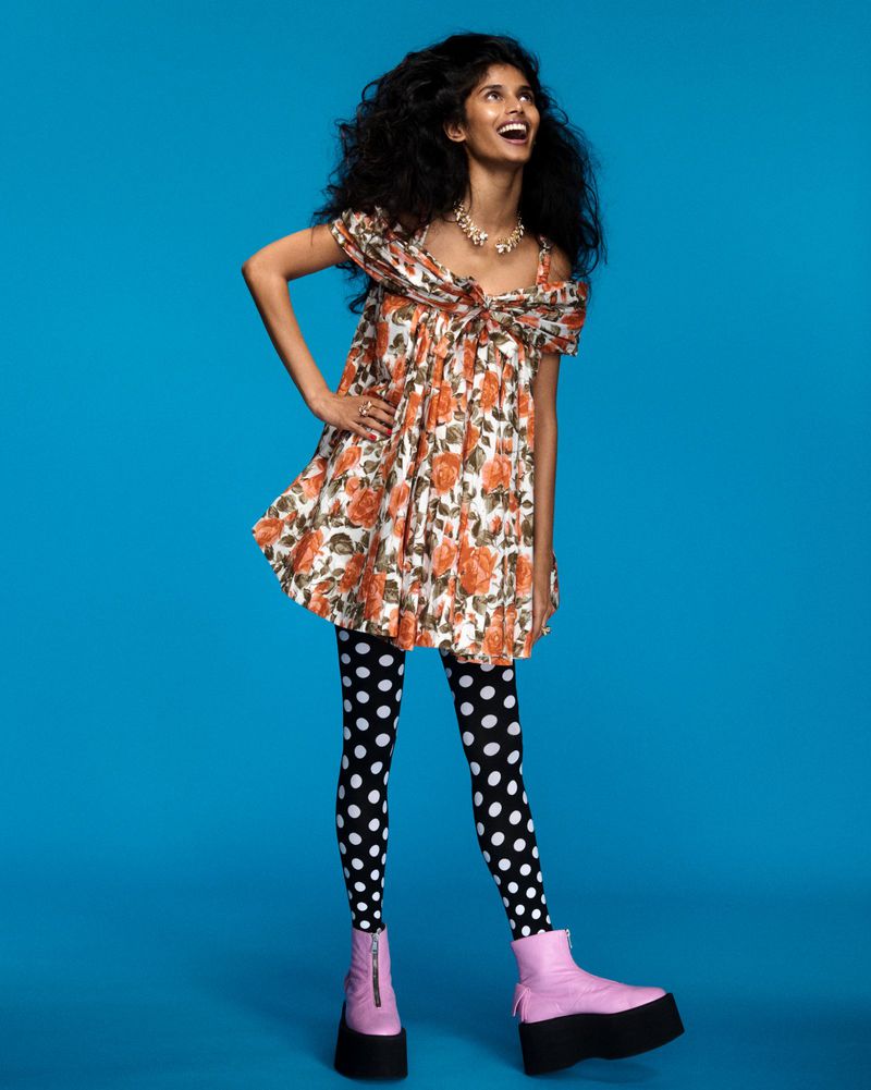 Smiling woman in a floral mini dress, polka dot leggings, and platform boots against a blue background. Studio shot, full-length.