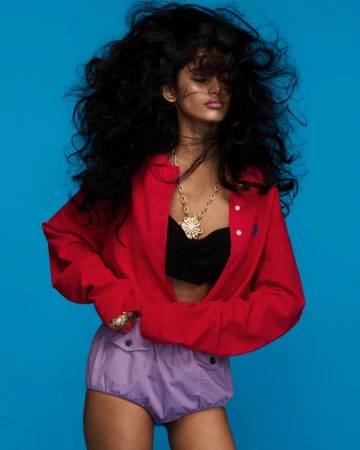 Fashionable woman with dark curly hair poses in a red shirt, black top, and lavender shorts against a blue background in a studio shot.