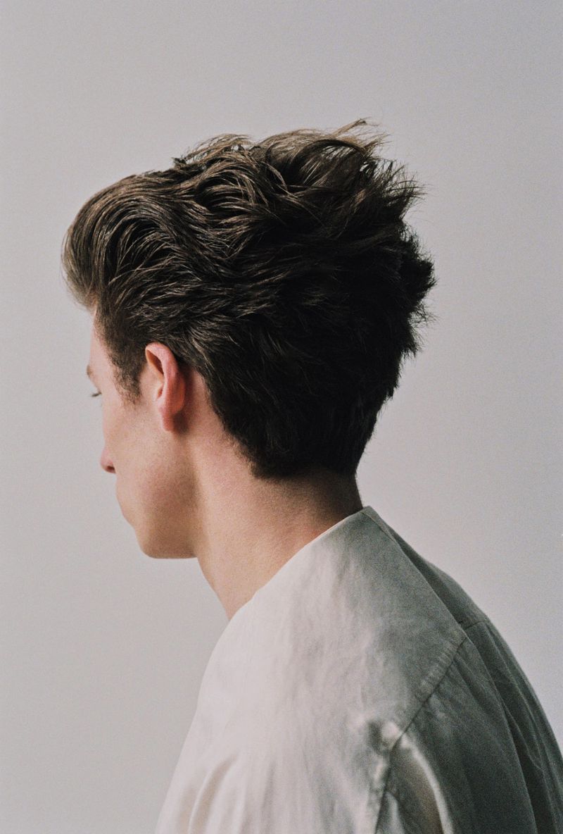 Profile view of a young adult male with short, dark wavy hair, wearing a casual light-colored shirt against a neutral background.