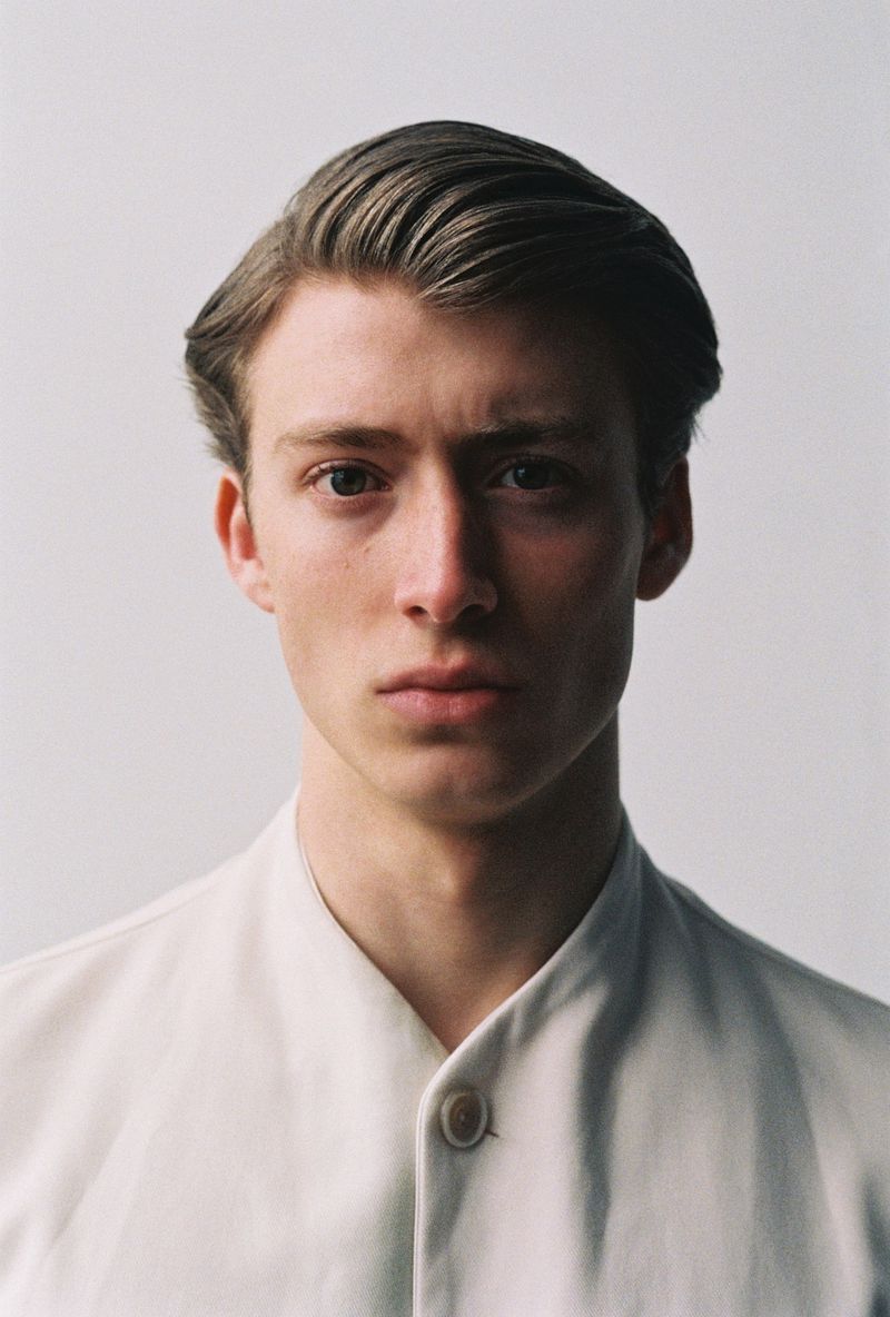 Portrait of a fair-skinned adult male with styled brown hair, wearing an off-white collared shirt against a light background in a studio.