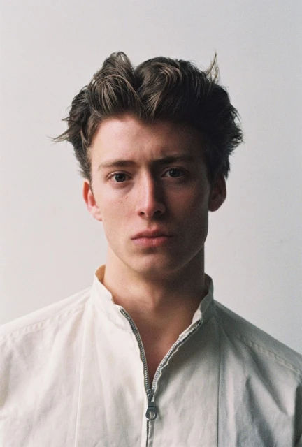 Close-up portrait of a young adult male with medium-length brown hair, wearing a white zip-up jacket, against a plain neutral background.