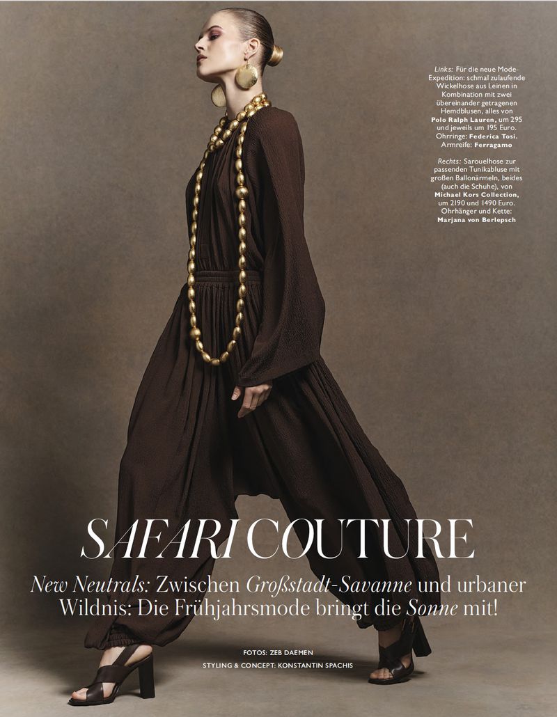 Fashion editorial shot of a woman in a brown jumpsuit, statement gold jewelry, and heeled sandals against a neutral brown background.