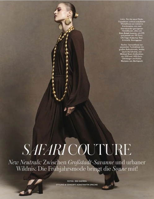 Fashion editorial shot of a woman in a brown jumpsuit, statement gold jewelry, and heeled sandals against a neutral brown background.