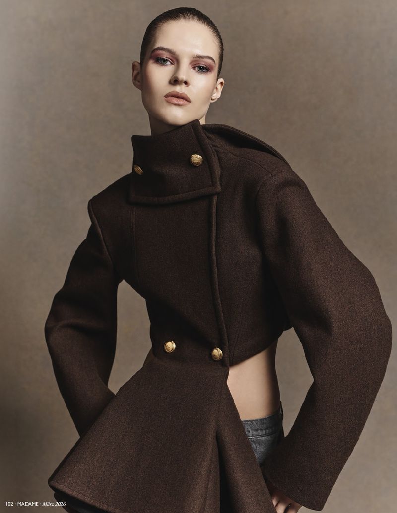 Fashion model in a studio setting wearing a brown cropped jacket with large gold buttons and grey denim jeans, arms slightly out.