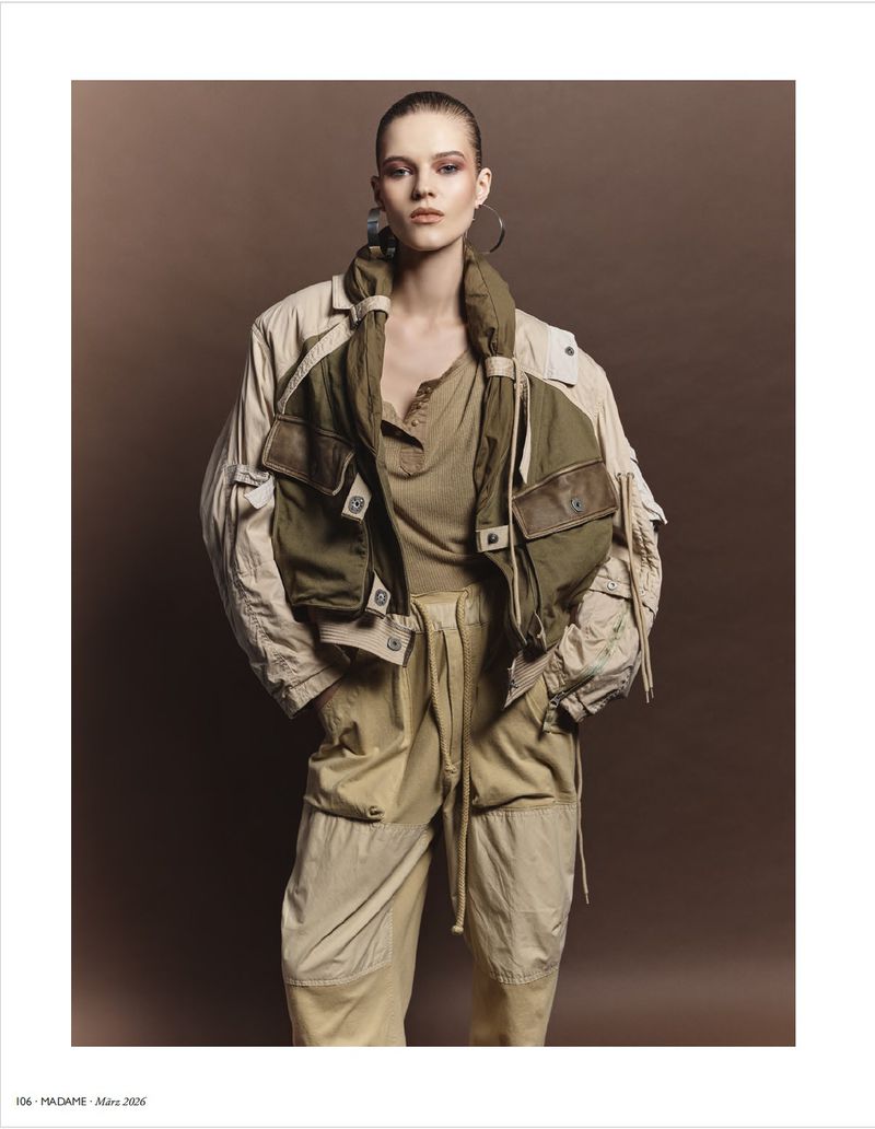 A fashion shot of a female model with slicked-back hair in a tan top, beige cargo pants, and a khaki jacket with large pockets against a brown background.