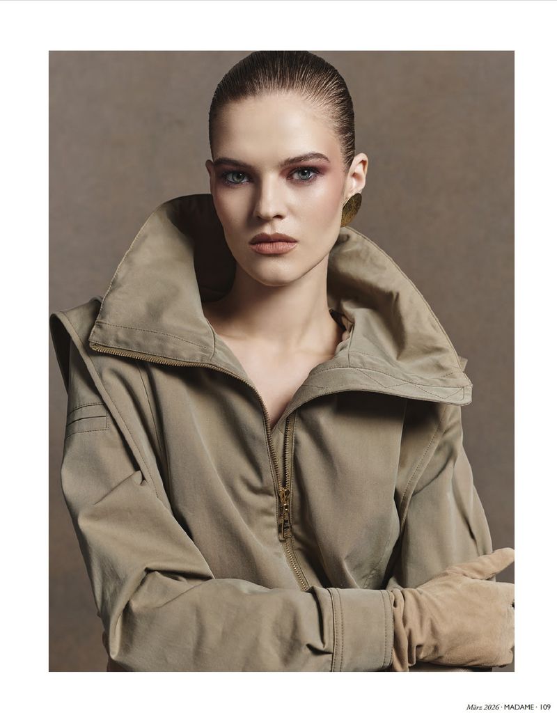 Close-up of a young woman with slicked-back hair in a khaki jacket and gloves, posing against a neutral gray backdrop in studio lighting.
