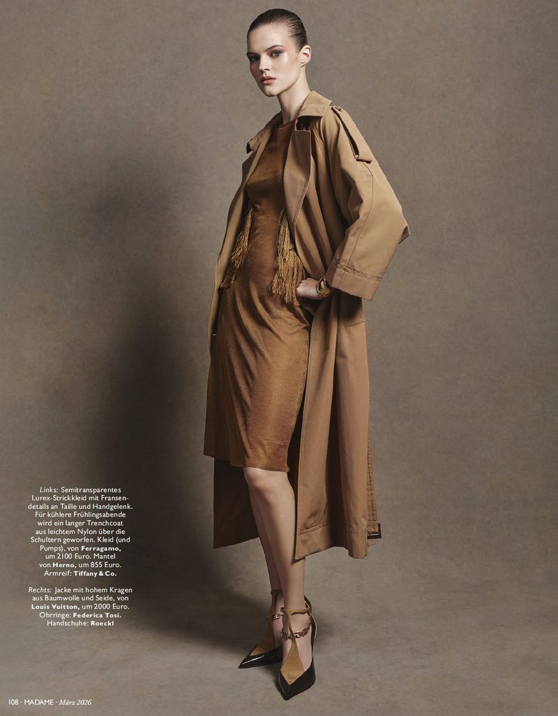 A woman in a brown dress and trench coat poses in a studio with gold heeled shoes, styled in a fashion editorial shot with neutral tones.