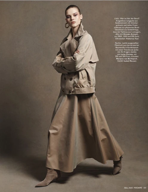 Fashion shot of a woman in a trench coat and maxi skirt. Model poses in an indoor studio setting with neutral background and soft lighting.