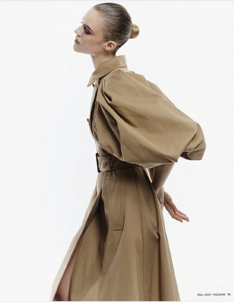 Model in profile wearing a belted camel coat with statement sleeves against a clean white background