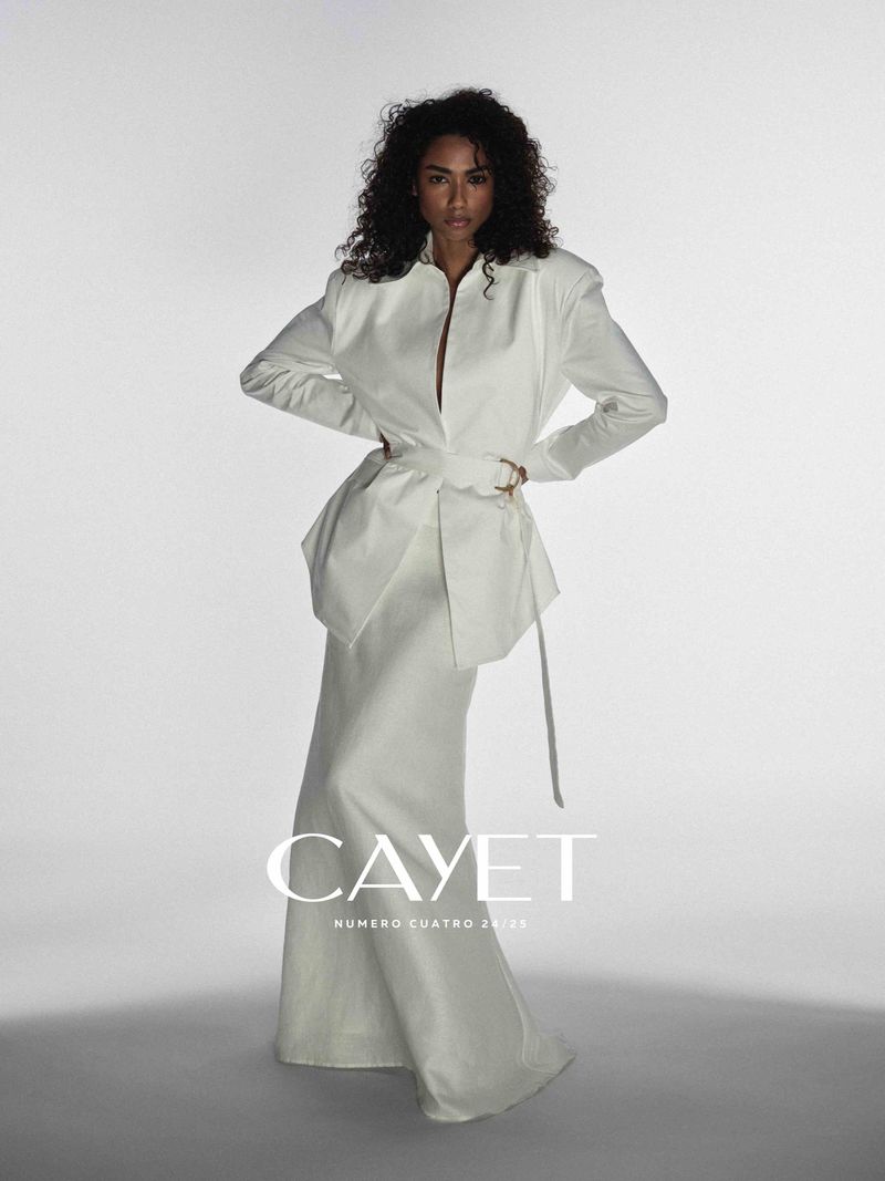Fashion editorial shot of a black female model posing in a long white dress with waist tie in a bright photo studio, arms on hips.
