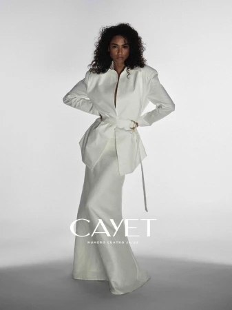 Fashion editorial shot of a black female model posing in a long white dress with waist tie in a bright photo studio, arms on hips.