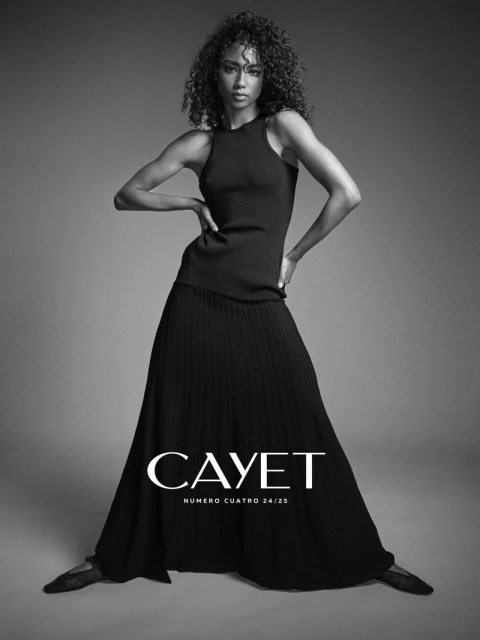 A slender Black woman poses in a sleeveless black top and black skirt with her hands on her hips in a studio shot.