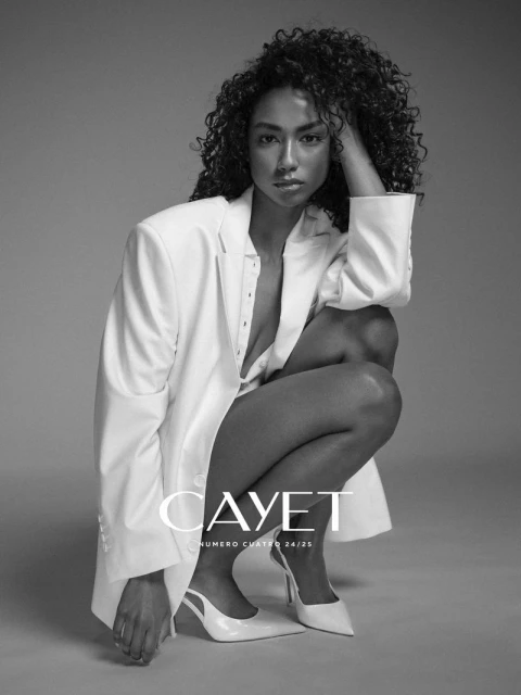 Fashion shot of a curly-haired woman in a white blazer and heels, squatting against a grey studio backdrop, showcasing beauty and style.