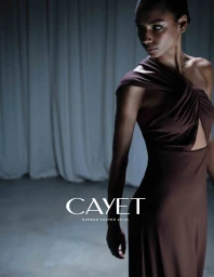 Model in a brown one-shoulder dress posing against draped white curtains in a low-lit studio setting for Cayet