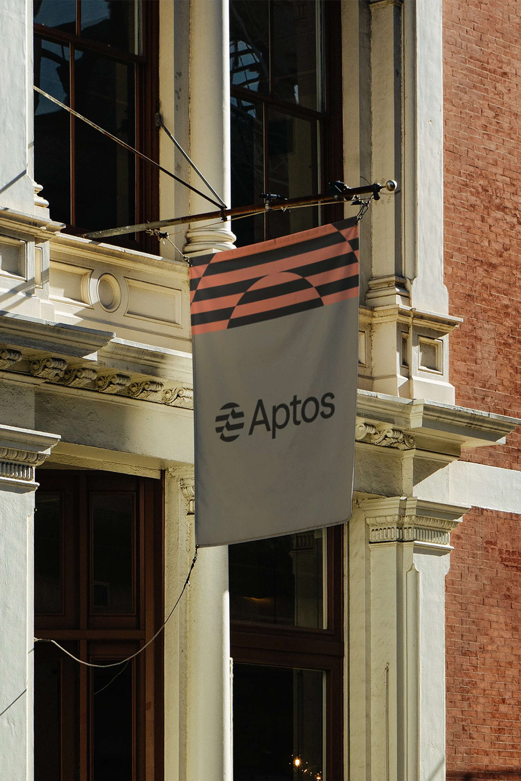 Aptos brand flag hanging on an ornate building with tall windows. The flag has a logo and horizontal lines pattern on top.