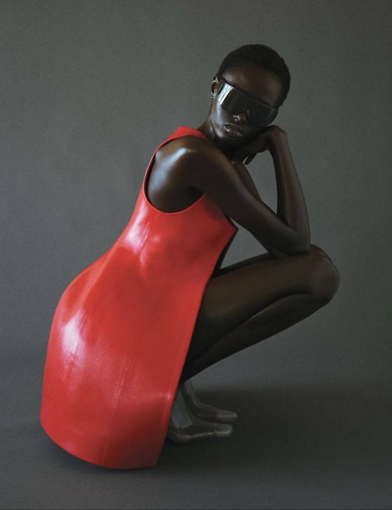 Fashion shot of a dark-skinned woman in a red dress and futuristic sunglasses, crouching against a grey backdrop in a studio setting.