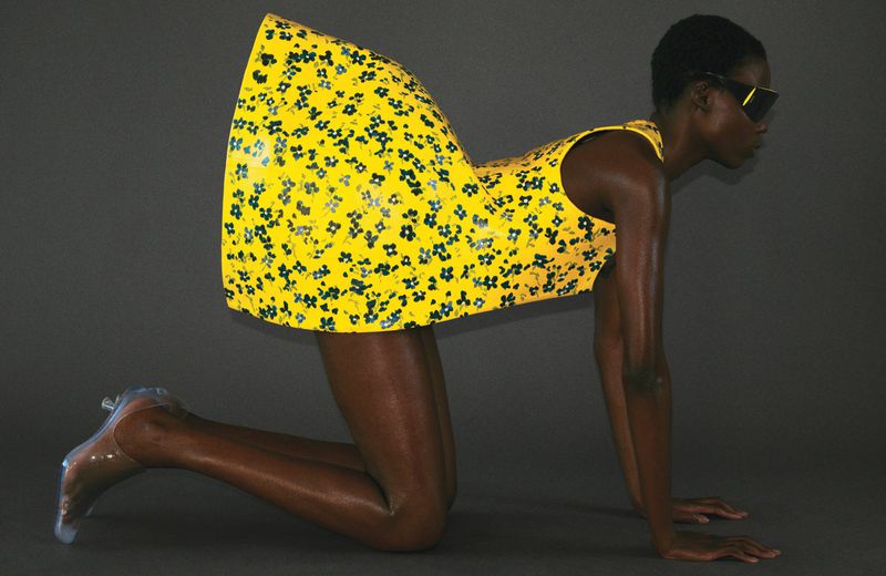 Fashion shot of an African woman on her hands and knees, wearing a bright yellow floral dress and transparent high heel shoes.