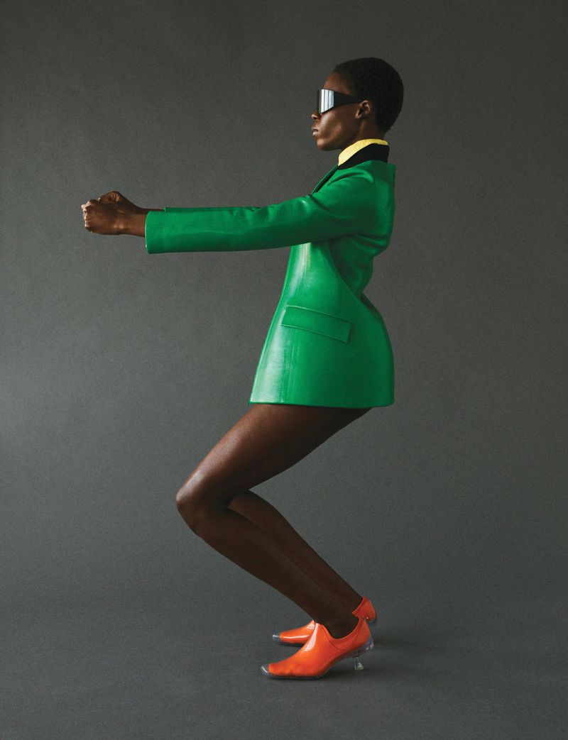 Profile shot of Black woman in green blazer suit and orange shoes, striking a playful pose against a neutral gray backdrop in studio.