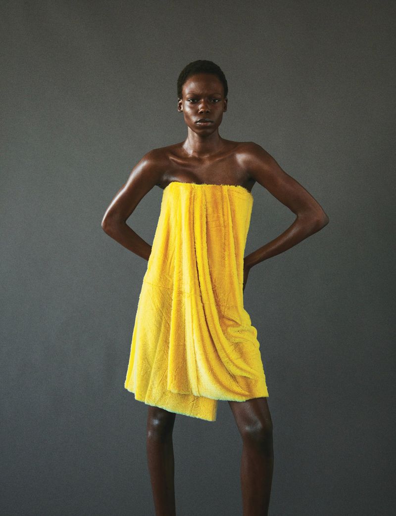 Elegant, dark-skinned woman strikes a fashionable pose in a draped, strapless, bright-yellow textile against a neutral backdrop.