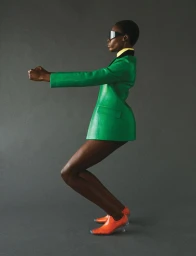 Model in a bright green blazer and orange shoes wearing sunglasses against a grey studio background