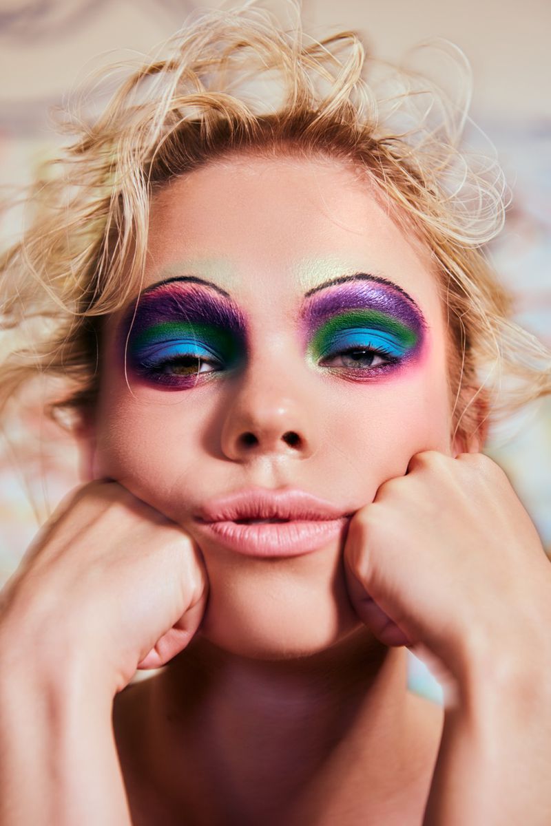 Close-up portrait of a young adult woman with heavily applied blue, green, and purple eyeshadow, holding her cheeks with both hands.