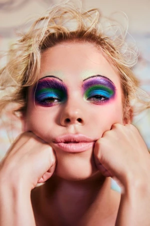 Close-up portrait of a young adult woman with heavily applied blue, green, and purple eyeshadow, holding her cheeks with both hands.