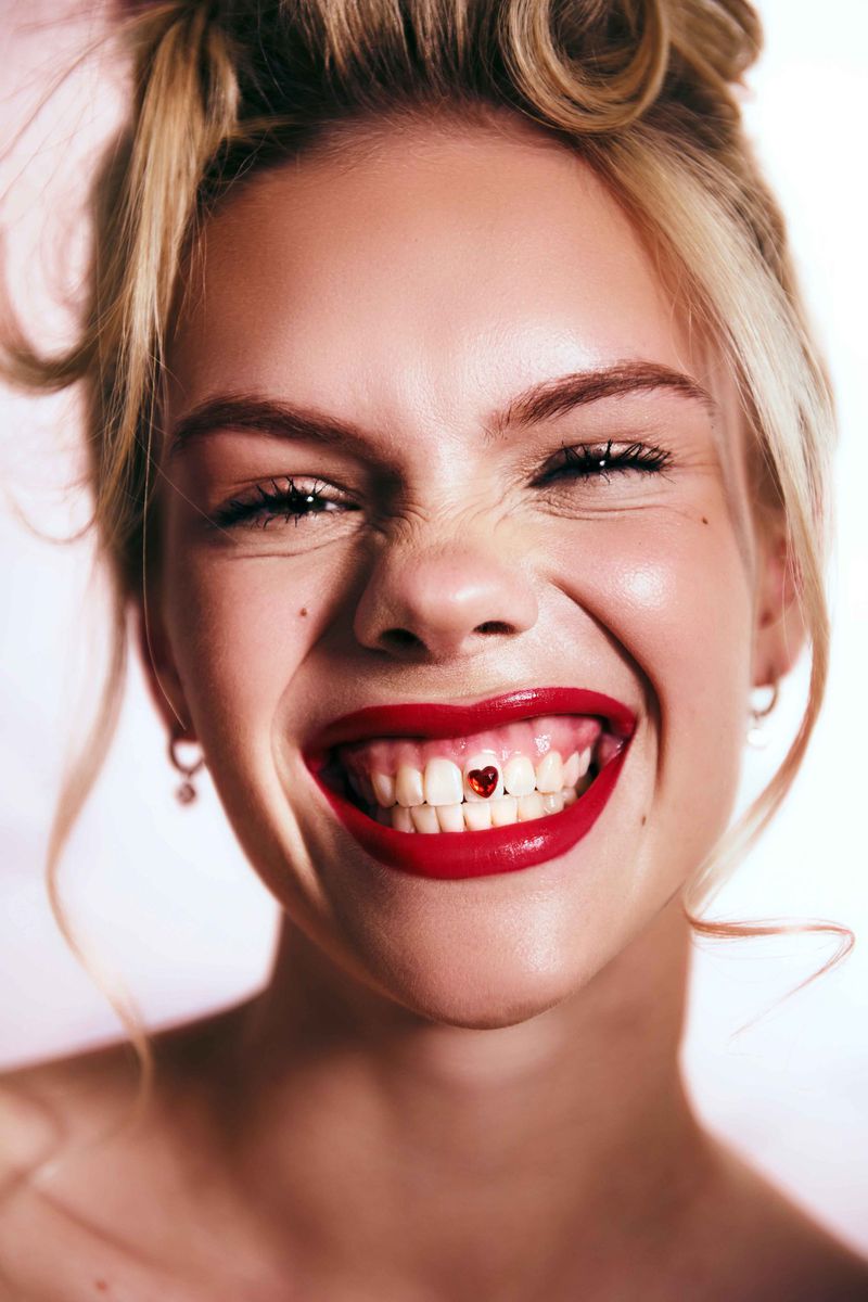 Close-up portrait of a fair-skinned young woman smiling wide, showcasing a red heart-shaped tooth gem, styled blonde hair, and red lipstick.