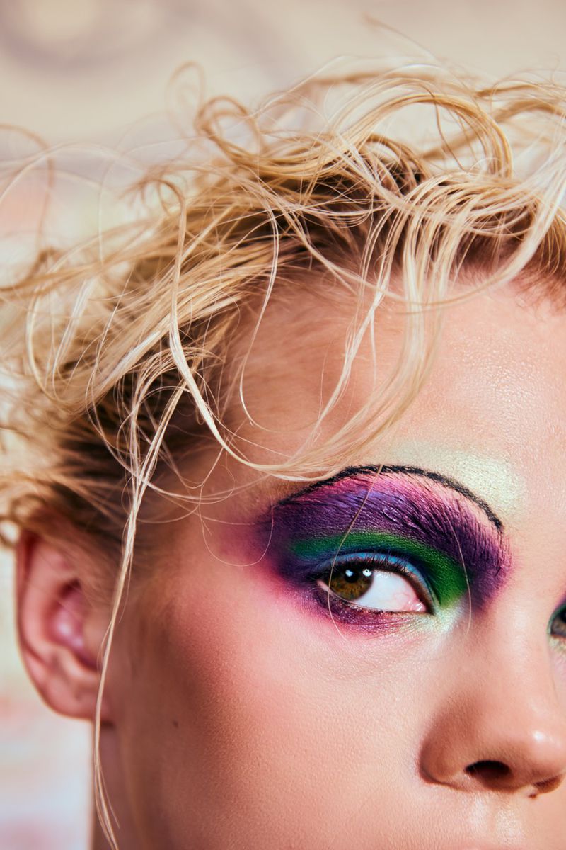 Close-up portrait of a woman's face featuring vibrant purple, green, and white eyeshadow, complemented by blonde hair and fair skin.