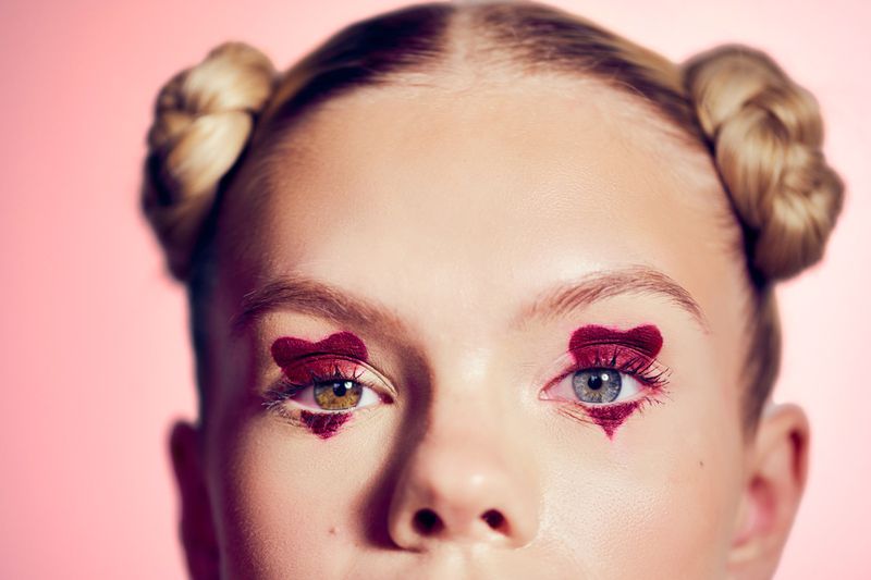 Close-up of a young woman's face with two-toned eyes, heart-shaped makeup around her eyes, and blonde hair in space buns, against a pink background.