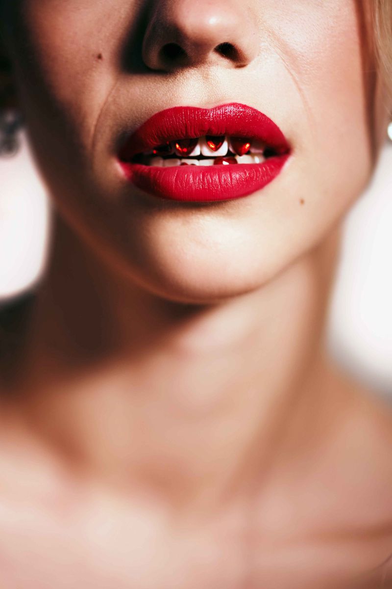Close-up of a woman's mouth with red lipstick and small red heart objects between teeth, focusing on beauty and unconventional style.
