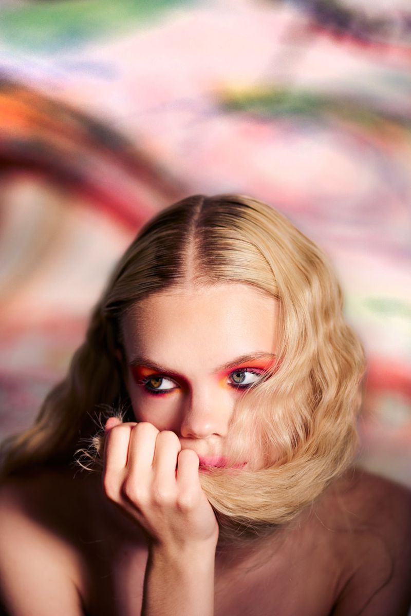 Close-up portrait of a young adult woman with blonde wavy hair, vibrant makeup, and a hand near her face, against an abstract colorful backdrop.