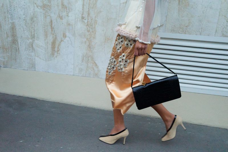 Fashionable woman in embroidered skirt and heels carrying a black purse walking along a sidewalk with a textured wall in background.