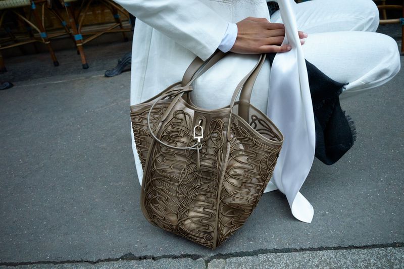 Person in white suit sitting next to a light brown leather patterned tote bag with silver hardware on concrete ground outdoors.