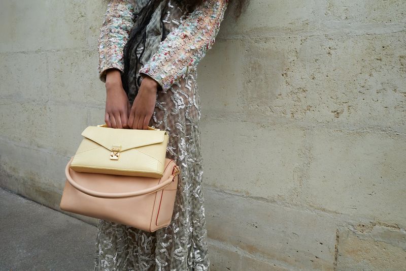 Person stands by wall, holding a yellow handbag atop a larger pink handbag, wearing a sparkling silver dress and black scarf.