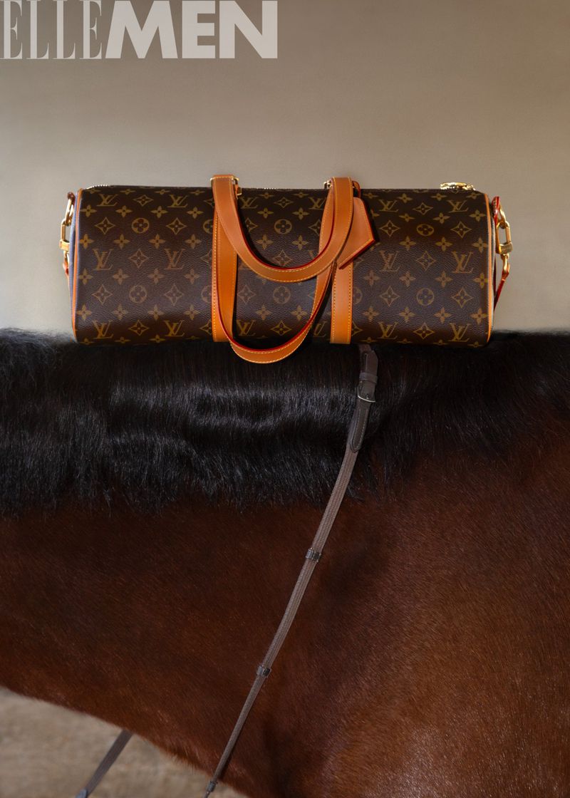 Louis Vuitton duffle bag on black horse back for Elle Men. Leather handles, logo pattern, indoor studio lighting, neutral background.