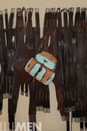 Aqua and brown satchel bag displayed against a background of dark leather straps with metal hardware hanging on a wall.