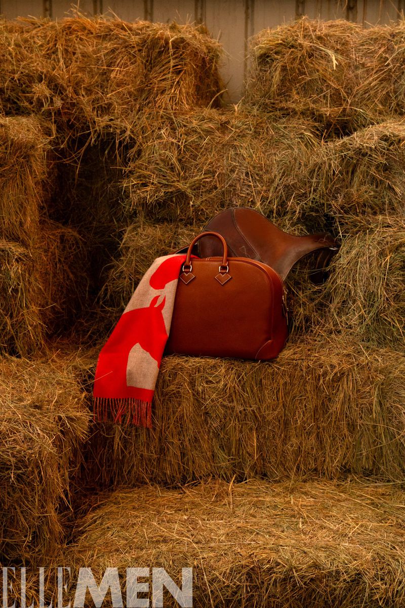 Leather duffel bag and horse-patterned scarf sit atop stacked hay bales in a rural setting, with 'ELLE MEN' logo in the corner.