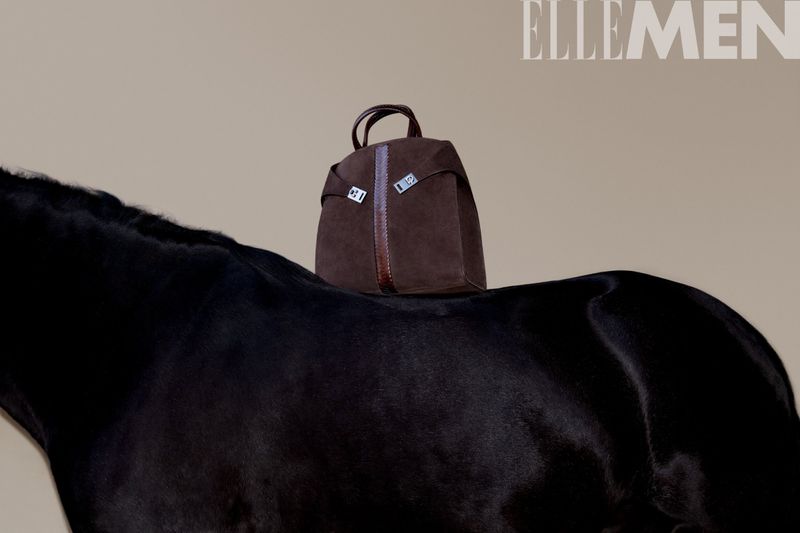 Brown leather bag sits atop a black horse against a neutral backdrop, photographed for Elle Men magazine, in a fashion product studio shot.
