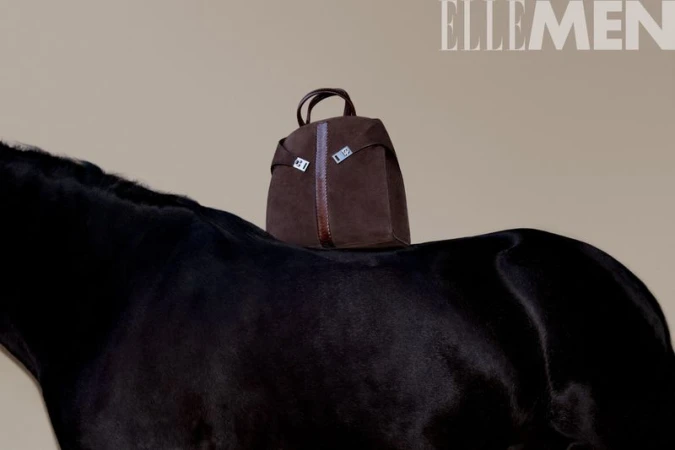 Brown leather bag sits atop a black horse against a neutral backdrop, photographed for Elle Men magazine, in a fashion product studio shot.