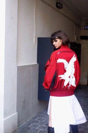 Woman in a red bomber jacket with an eagle design and a white skirt stands in front of a concrete building in an urban setting.