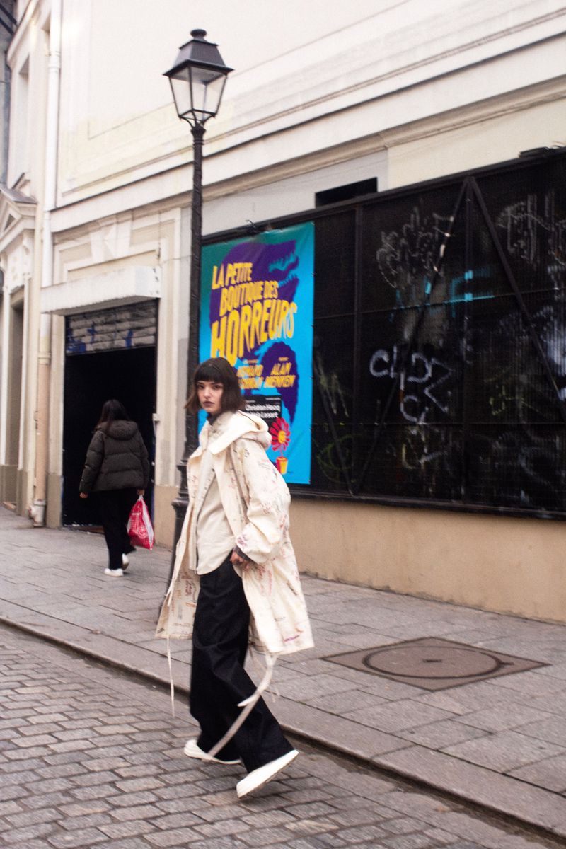 A fashionable adult female strides down a cobblestone street in a loose coat, black pants, and white shoes with building and poster background.