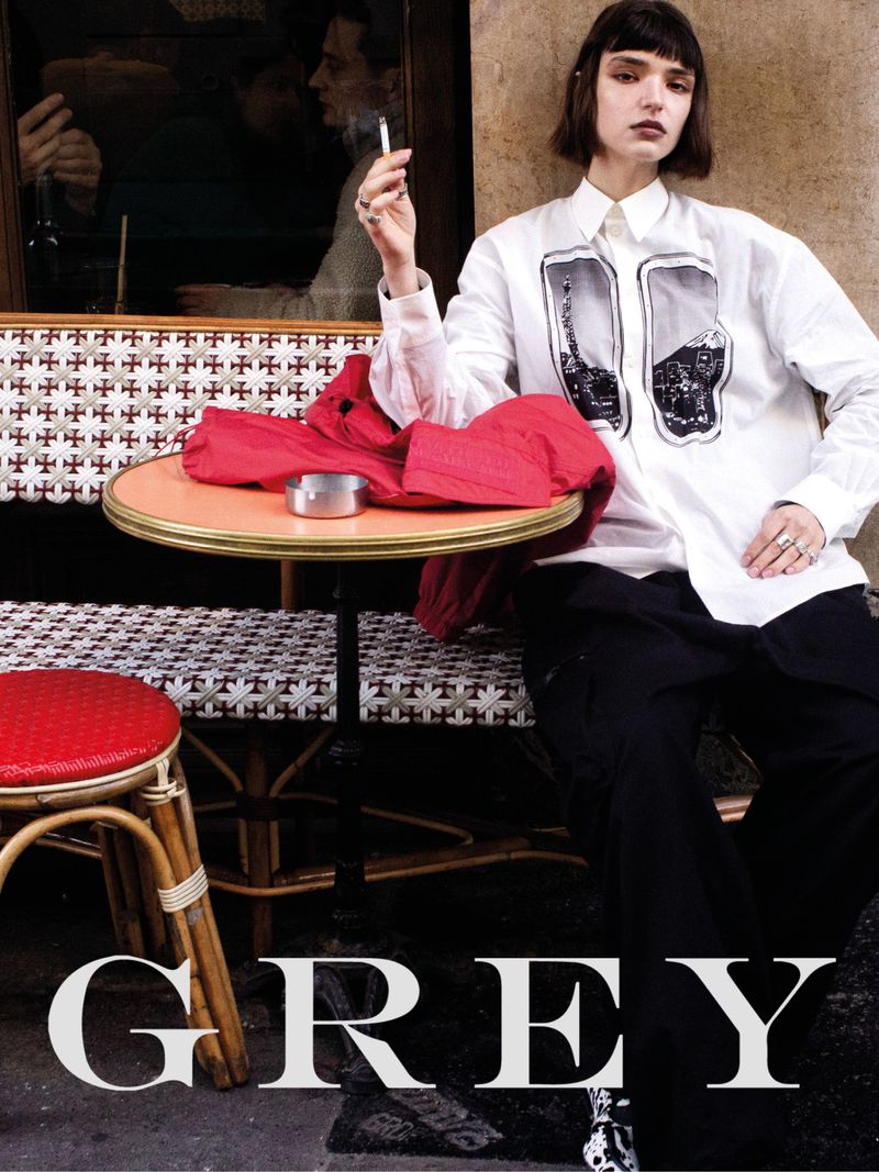 Fashionable young woman in a white shirt with an Eiffel Tower graphic, poses near a café table, smoking a cigarette in an urban setting.