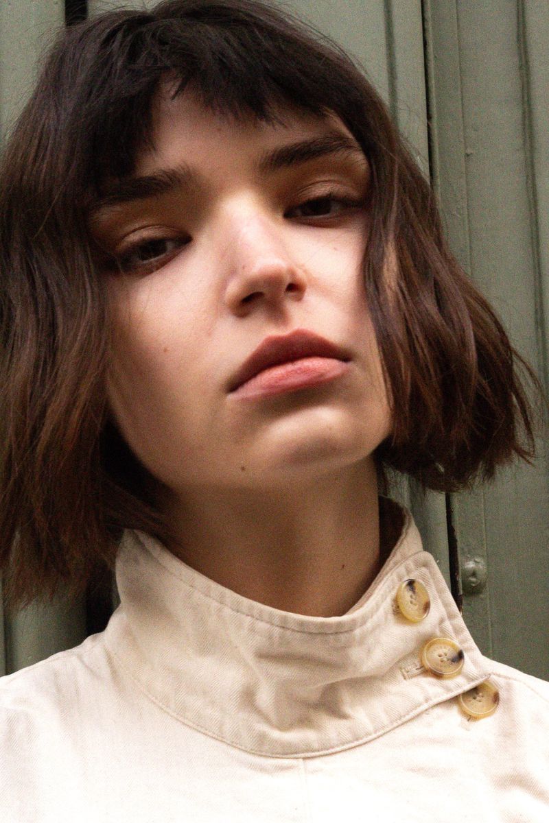 Portrait of a young brunette woman with bangs wearing a beige buttoned shirt in front of a muted green wall, captured in natural light.