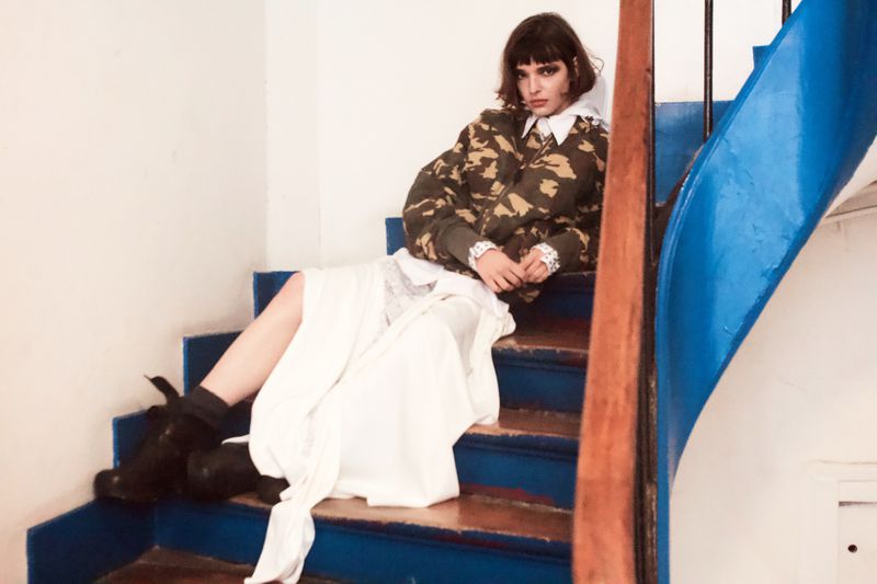 A model sits on a blue staircase wearing a white dress and a camouflage print top, looking directly at the camera indoors.