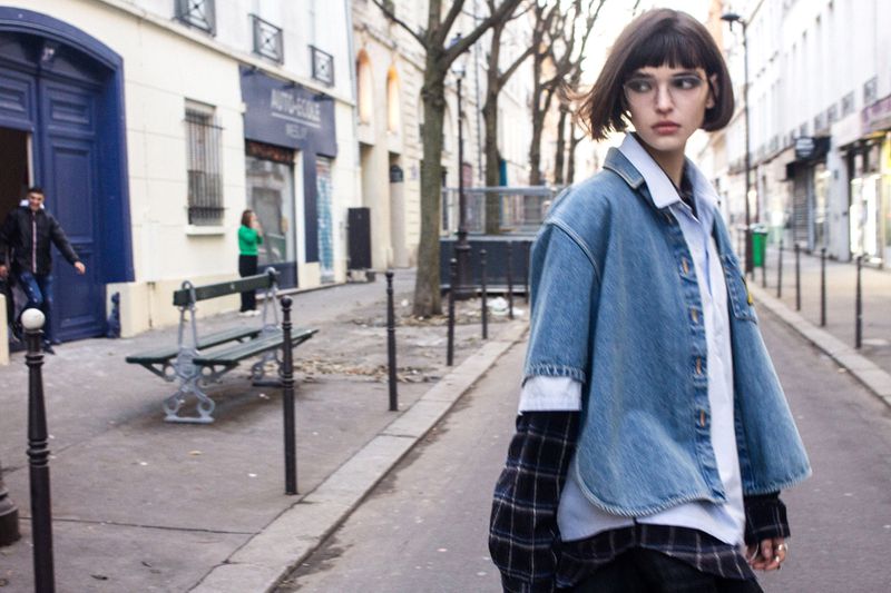 A young adult woman poses in an urban setting, wearing a denim jacket over a layered shirt, with buildings and a street in the background.