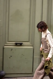 Woman walking past a large green door wearing striped overalls and a green sweater tied around her waist.