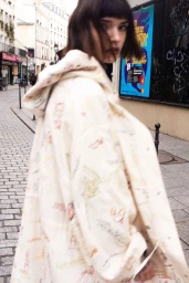 Model with dark hair wearing a patterned cream hooded coat walking down a cobblestone European street.