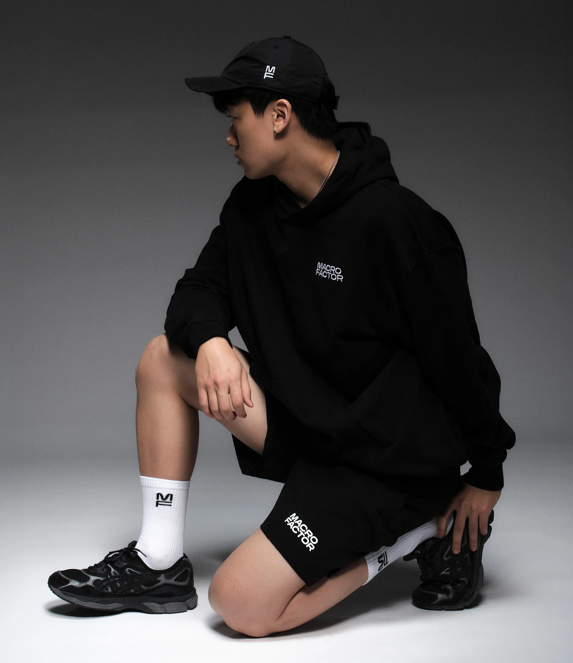 Asian man in a black hoodie and shorts kneeling in a photo studio. He wears a black baseball cap and black trainers. Plain gray background.