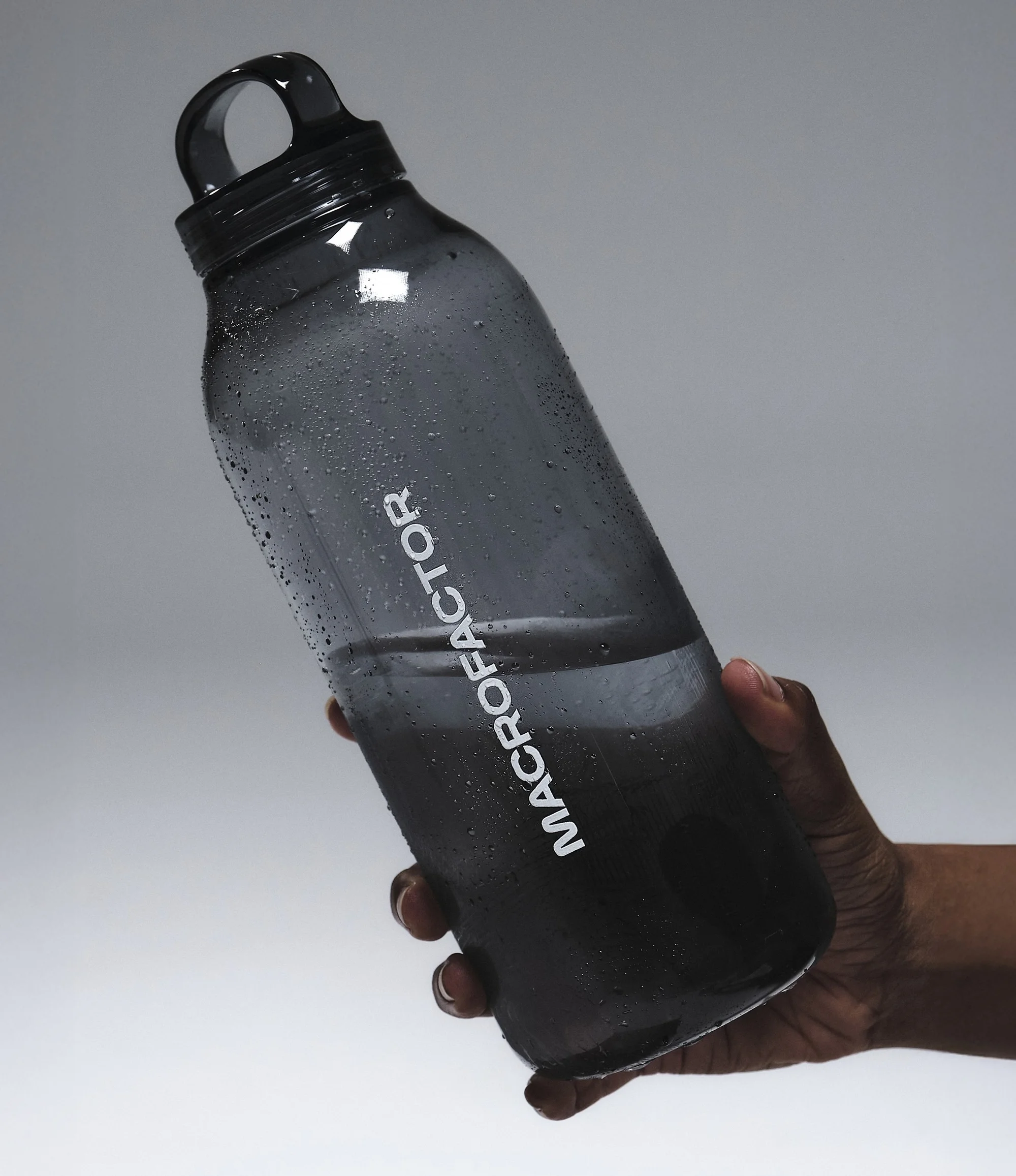 Person holding a translucent grey MacroFactor-branded water bottle with water droplets, against a graduated grey background.
